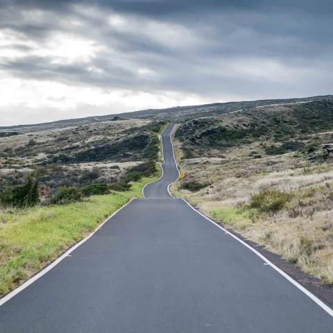 A narrow road winding through rolling hills under a cloudy sky.