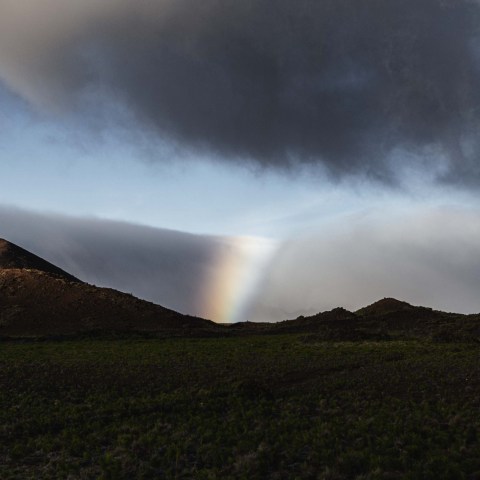Dramatic landscape with hills, dark clouds, and partial rainbow in the distance.